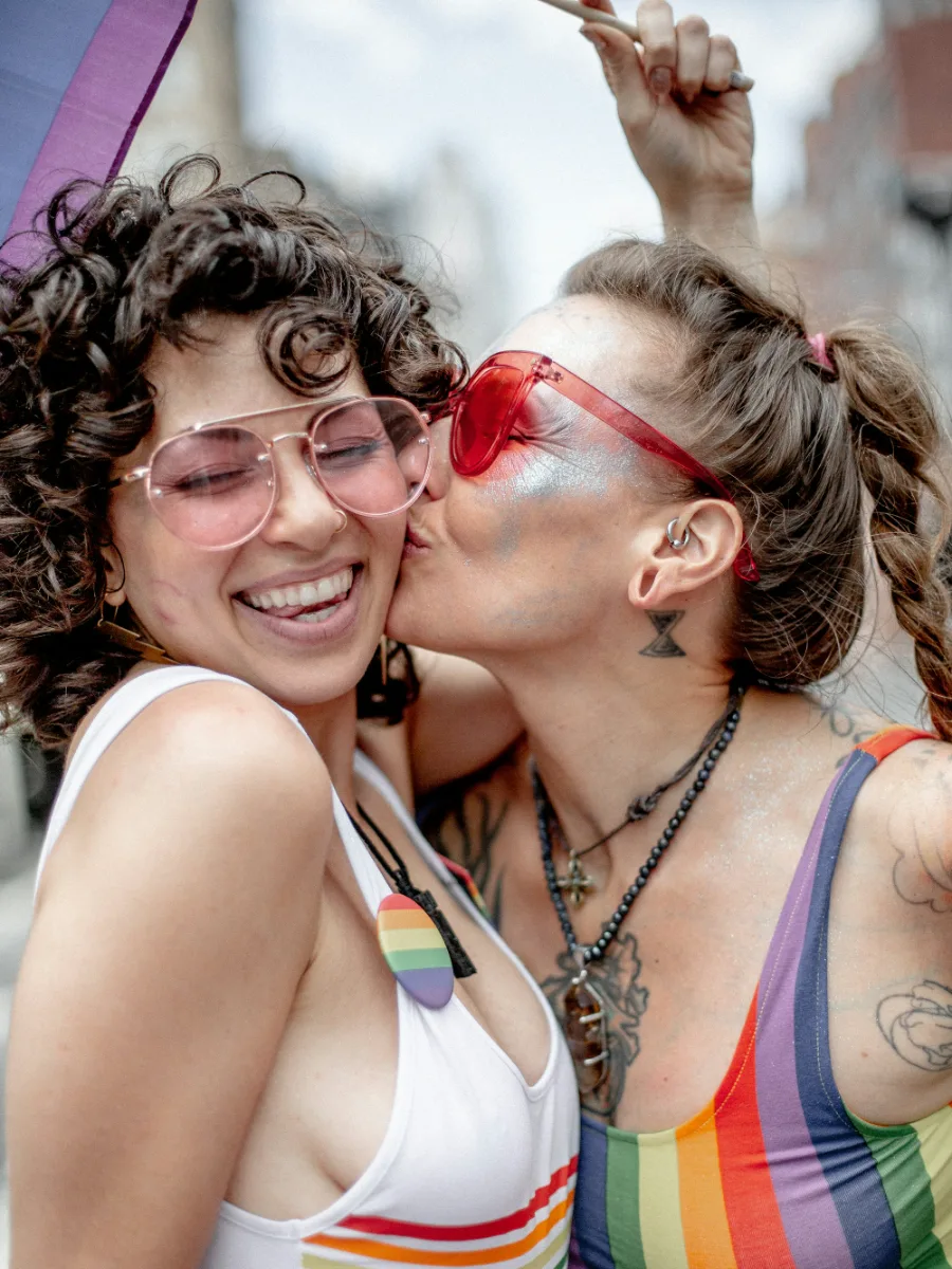 Deux femmes rieuses à la Pride, l’une embrasse l’autre sur la joue